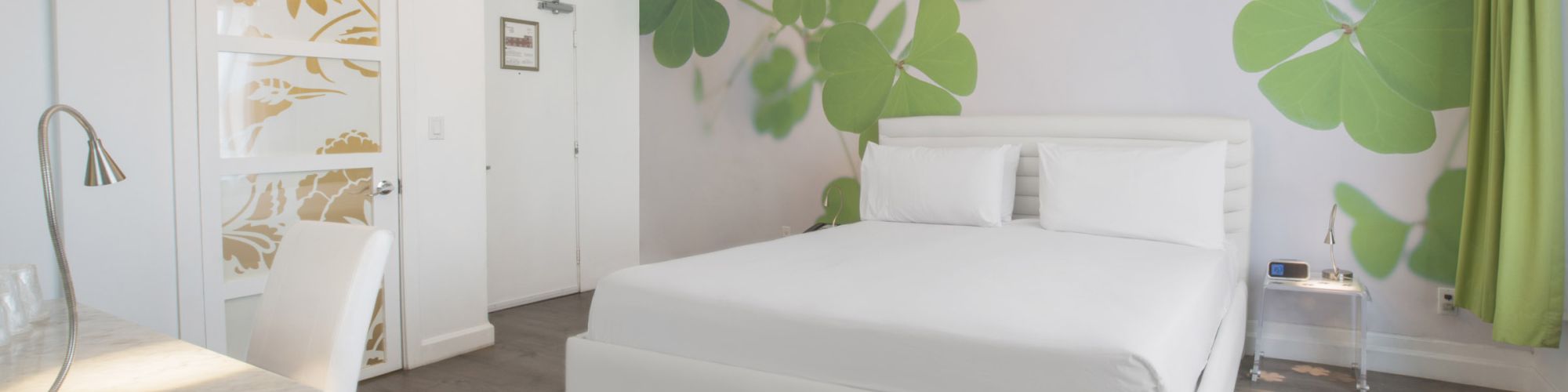 A modern bedroom with a white bed, leaf-patterned accent wall, chandelier, and green curtains.