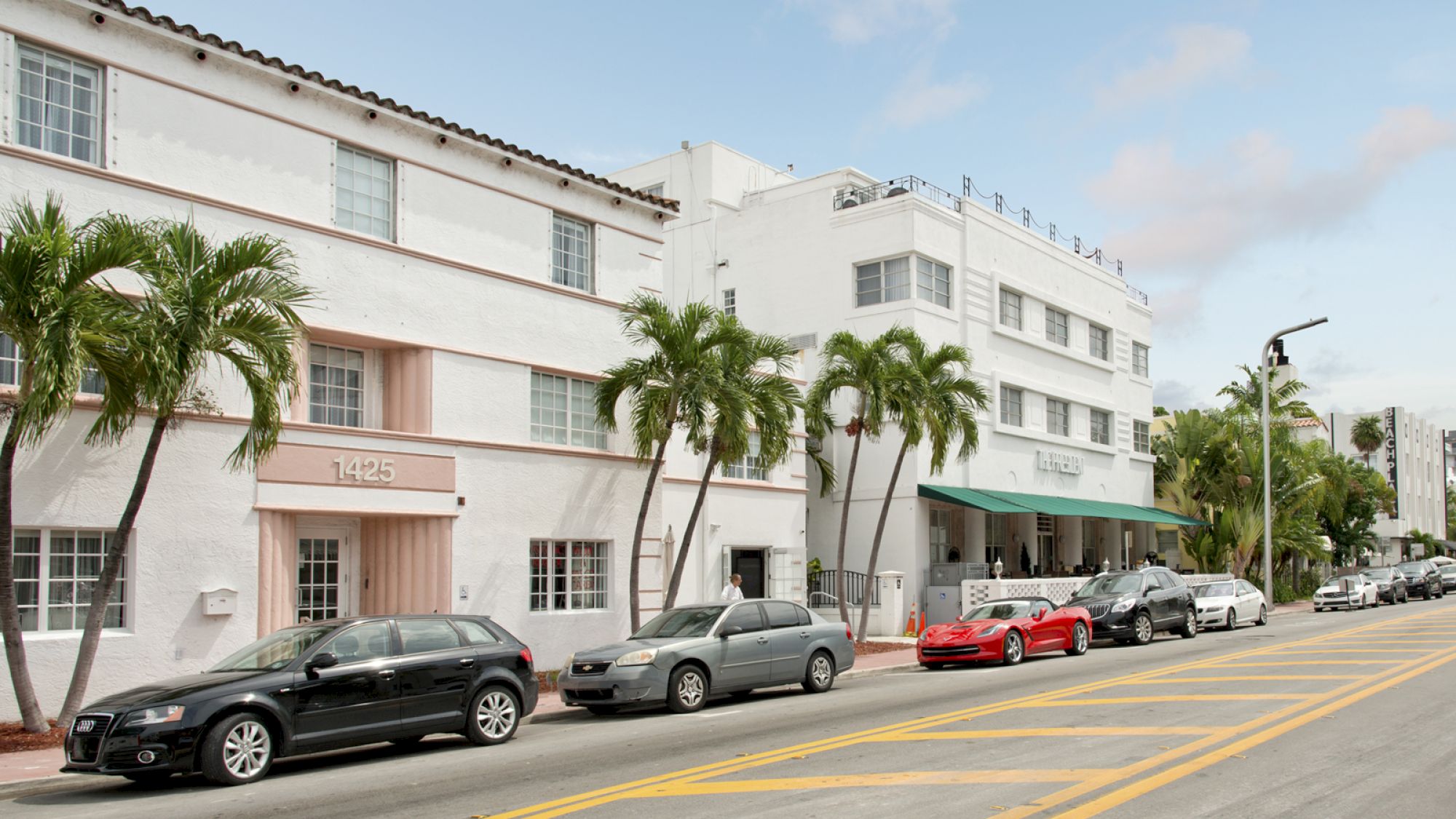 A row of white, multi-story buildings with palm trees, several parked cars, and a street with visible yellow lines on an overcast day.