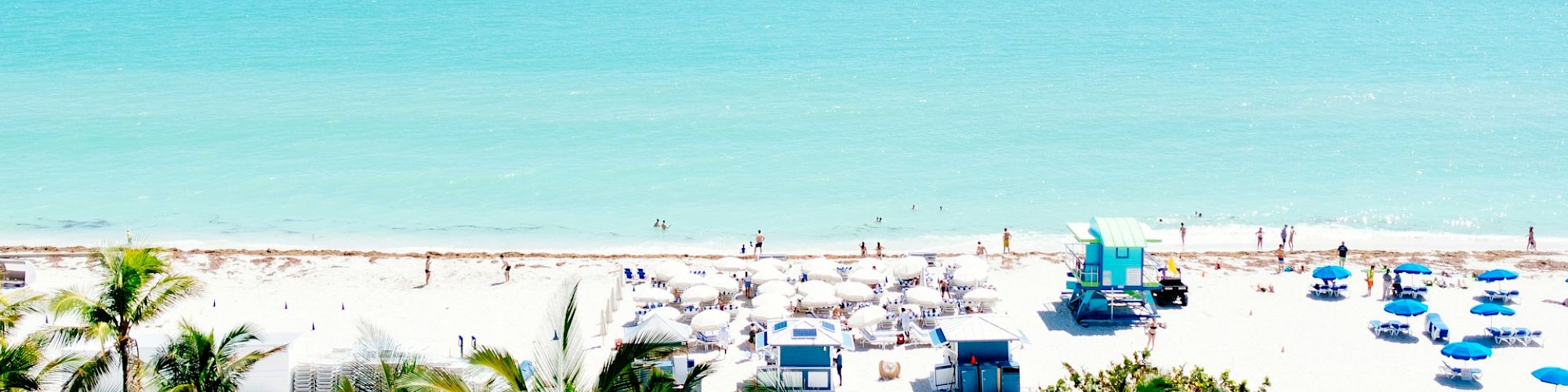 A sunny beach scene with turquoise water, white sand, palm trees, beach chairs, and umbrellas. Sparse cloud cover and a few people present.