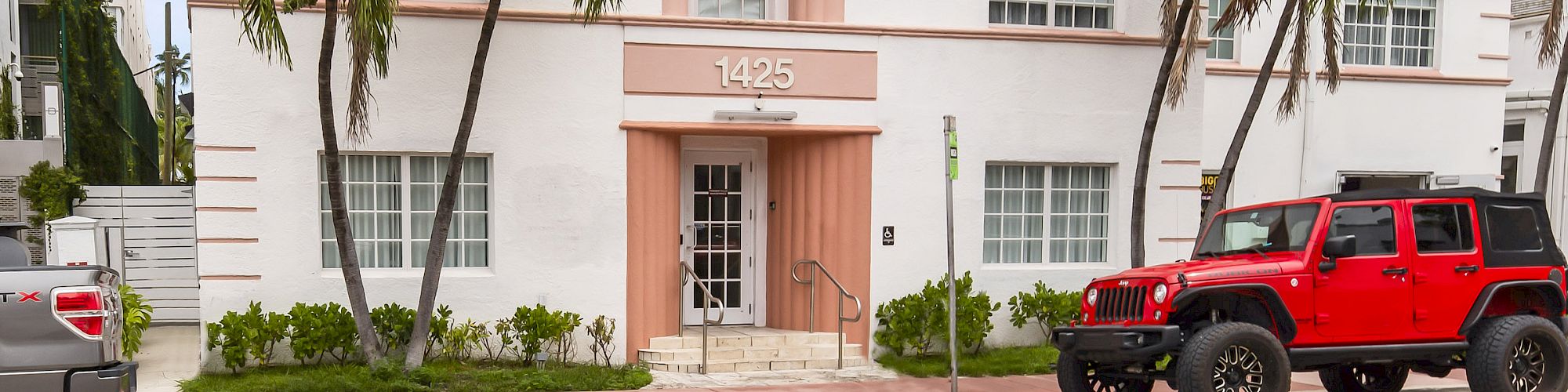 A three-story white building with pink accents, palm trees, and parked vehicles in front, including a red Jeep, under a blue sky.