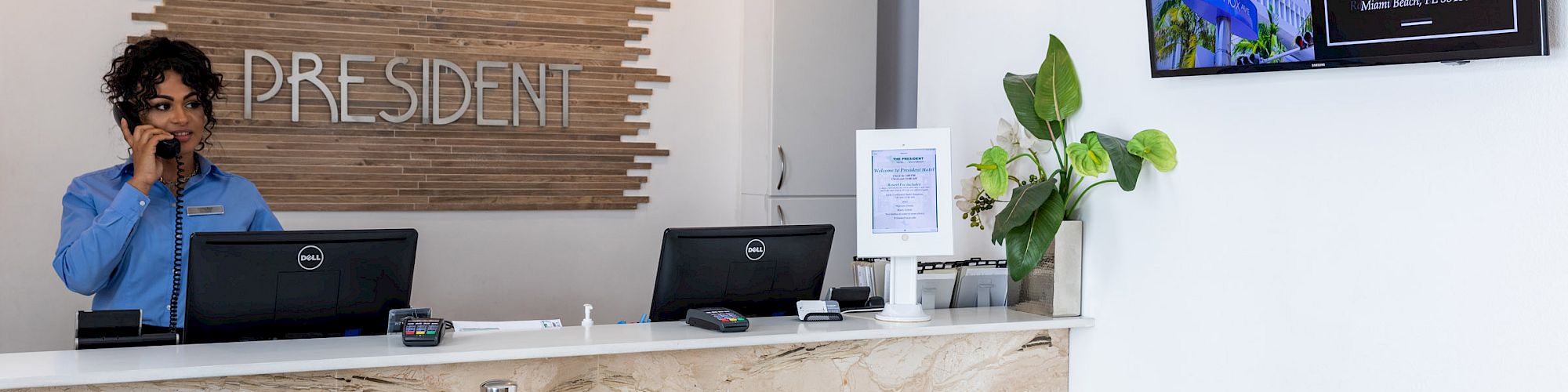 A receptionist stands at a counter with two computers, a plant, and a wall-mounted screen showing 