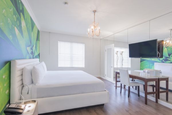 A modern bedroom with a large bed, wall art, chandelier, wall-mounted TV, desk, chair, and a mirrored wall enhancing the spacious feel.