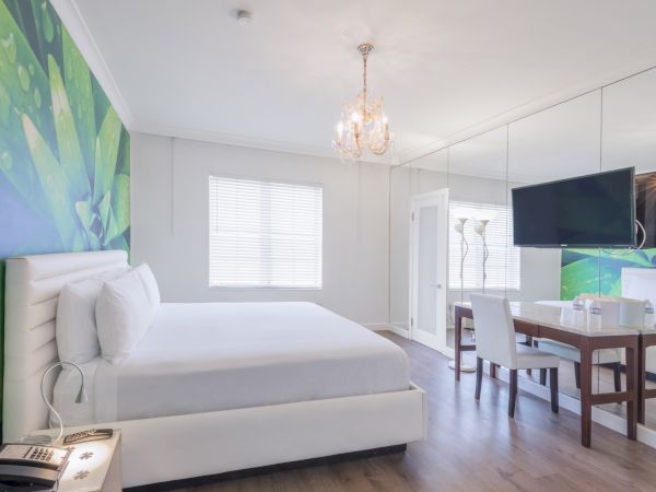 A modern bedroom with a large bed, wall art, chandelier, wall-mounted TV, desk, chair, and a mirrored wall enhancing the spacious feel.