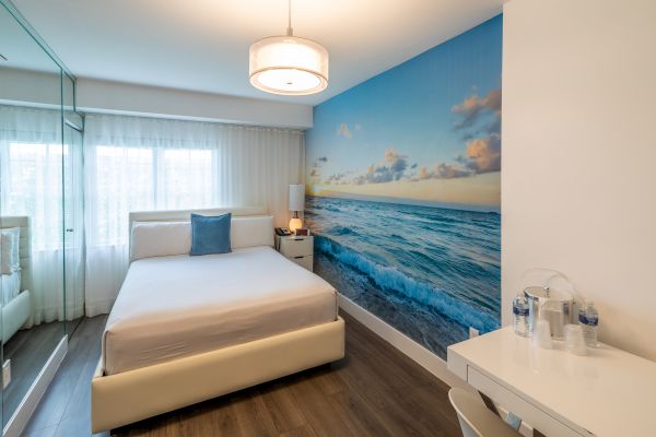 A modern bedroom with a beach-themed mural on the wall, a bed, nightstand, and a desk with bottled water, under a ceiling light fixture.