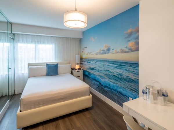 A modern bedroom with a beach-themed mural on the wall, a bed, nightstand, and a desk with bottled water, under a ceiling light fixture.