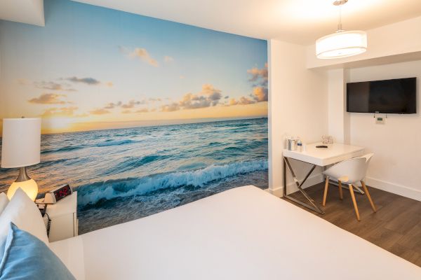 A modern hotel room features a large ocean mural, a white bed, a desk, a chair, and a wall-mounted TV.