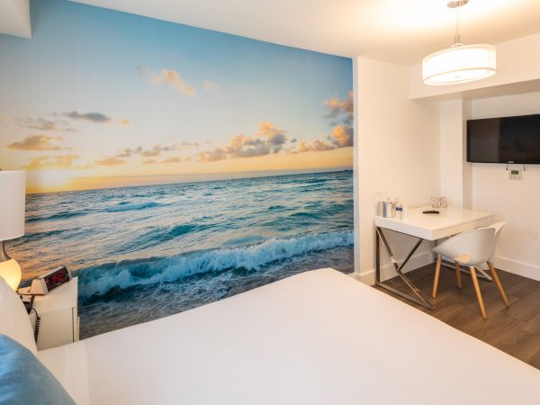 A modern hotel room features a large ocean mural, a white bed, a desk, a chair, and a wall-mounted TV.