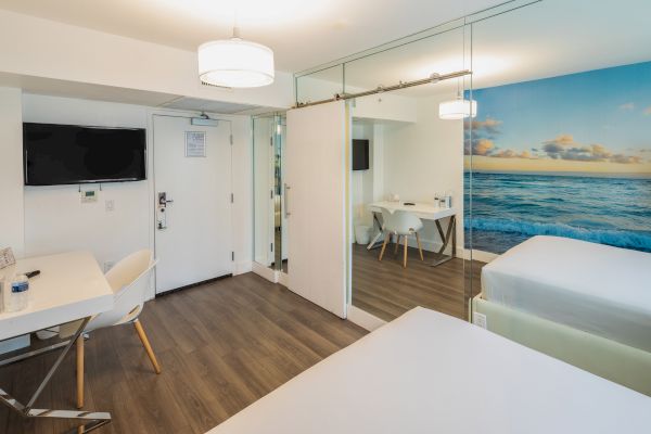 The image shows a modern hotel room with two beds, a desk, a TV, a dining area, and an ocean-themed wall mural.