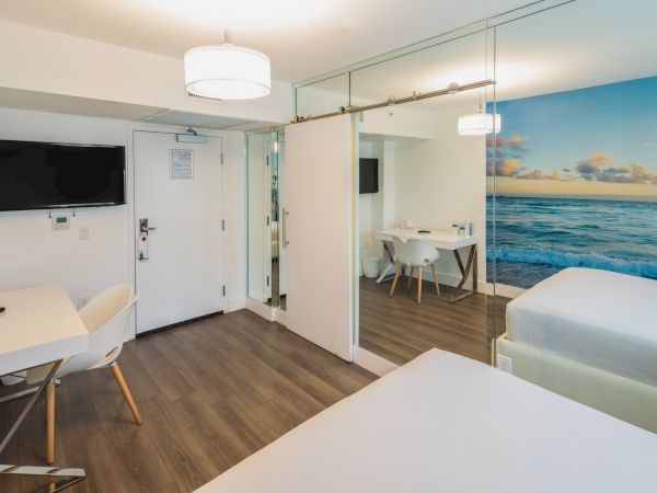 The image shows a modern hotel room with two beds, a desk, a TV, a dining area, and an ocean-themed wall mural.