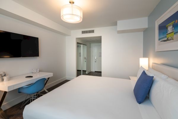 A modern hotel room features a bed with blue accents, a desk with a chair, a wall-mounted TV, and a doorway leading to another area.