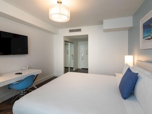 A modern hotel room features a bed with blue accents, a desk with a chair, a wall-mounted TV, and a doorway leading to another area.