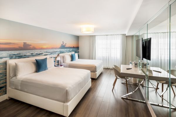 The image shows a modern hotel room with two beds, a large ocean-themed mural, a desk, chairs, and a TV, featuring wooden floors.