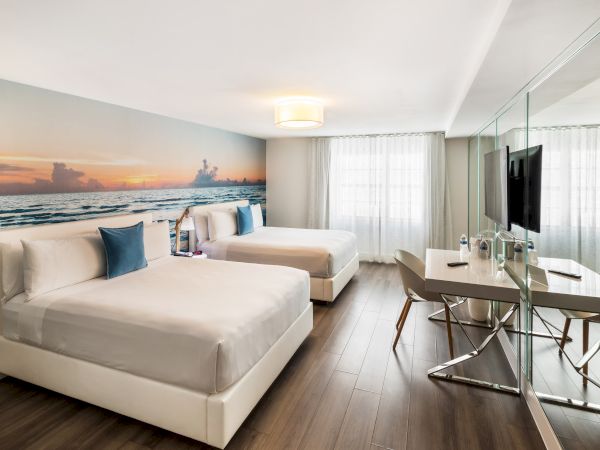 The image shows a modern hotel room with two beds, a large ocean-themed mural, a desk, chairs, and a TV, featuring wooden floors.