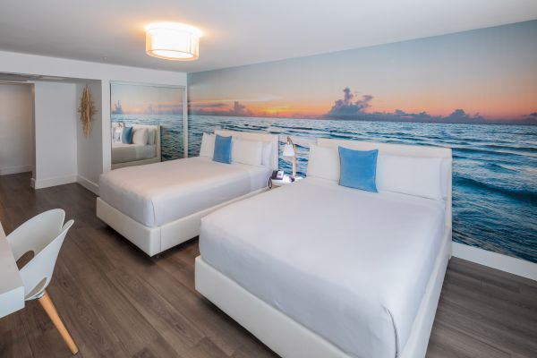 A modern hotel room with two beds, ocean mural, white bedding, blue pillows, wooden floor, and a light fixture above completes the scene.