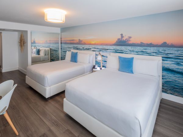 A modern hotel room with two beds, ocean mural, white bedding, blue pillows, wooden floor, and a light fixture above completes the scene.
