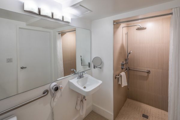 A modern bathroom with a sink, mirror, and accessible shower area equipped with grab bars and a handheld showerhead, featuring neutral tones.