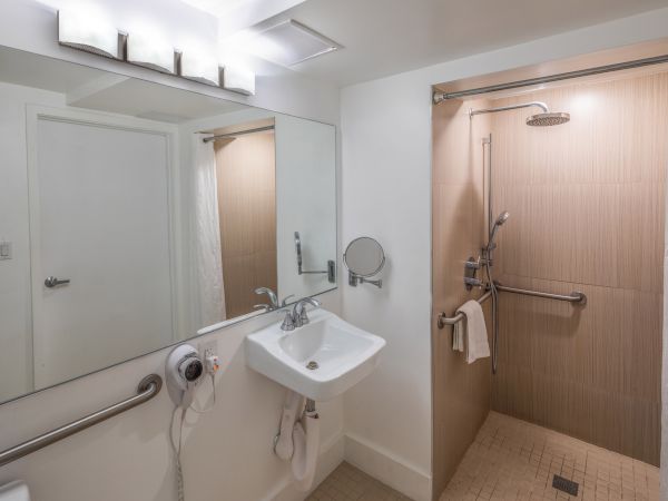 A modern bathroom with a sink, mirror, and accessible shower area equipped with grab bars and a handheld showerhead, featuring neutral tones.