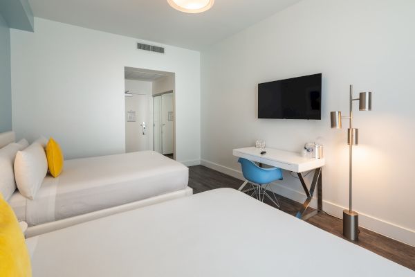 The image shows a hotel room with two beds, a desk, a chair, a TV on the wall, and a floor lamp, featuring modern and minimalist decor.