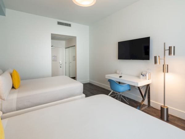 The image shows a hotel room with two beds, a desk, a chair, a TV on the wall, and a floor lamp, featuring modern and minimalist decor.