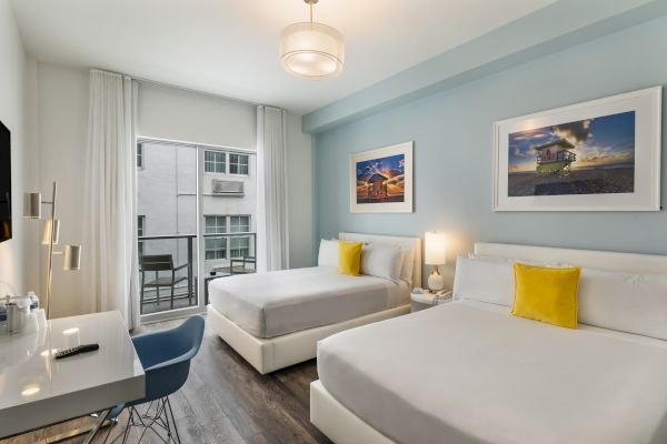 A modern hotel room with two beds, blue accent wall, framed pictures, a work desk, chair, and a small balcony with a view.