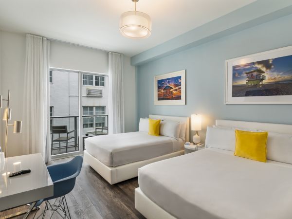 A modern hotel room with two beds, blue accent wall, framed pictures, a work desk, chair, and a small balcony with a view.
