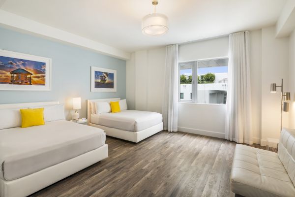 A modern bedroom with two beds, wood flooring, framed art, a window, and a small couch, decorated in light colors with yellow accents.