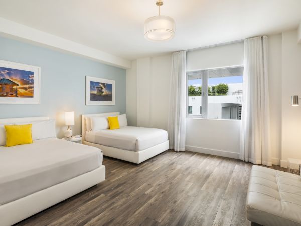 A modern bedroom with two beds, wood flooring, framed art, a window, and a small couch, decorated in light colors with yellow accents.