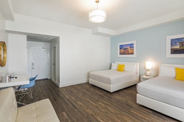 A modern hotel room with two beds, a small sitting area, and a desk. Wood flooring complements white walls and minimalist decor.