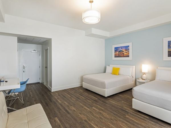 A modern hotel room with two beds, a small sitting area, and a desk. Wood flooring complements white walls and minimalist decor.