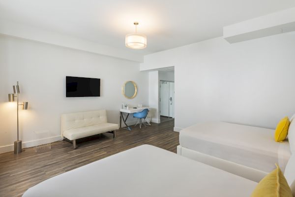 A modern hotel room with two beds, a wall-mounted TV, beige couch, desk with a mirror, and minimalist decor and lighting.