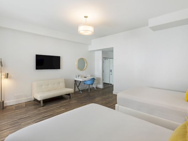 A modern hotel room with two beds, a wall-mounted TV, beige couch, desk with a mirror, and minimalist decor and lighting.