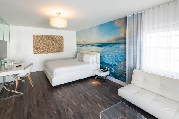 A modern hotel room with a bed, desk, sofa, sea-themed mural, and wood flooring. Bright and minimalist design is featured in the space.