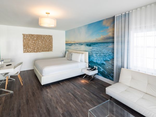 A modern hotel room with a bed, desk, sofa, sea-themed mural, and wood flooring. Bright and minimalist design is featured in the space.