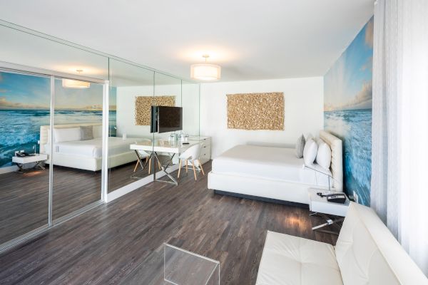 A modern bedroom with ocean-themed wallpaper, white furniture, mirrored closet doors, and a small desk with a TV.