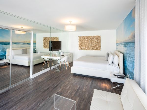 A modern bedroom with ocean-themed wallpaper, white furniture, mirrored closet doors, and a small desk with a TV.