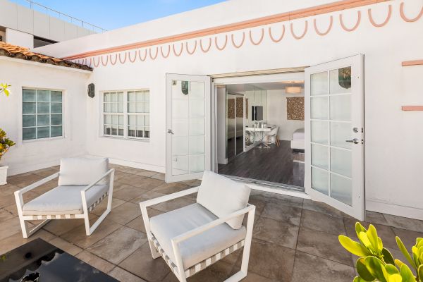 A sunlit patio features two white chairs, leading into a bright room through open double doors in a modern white building.