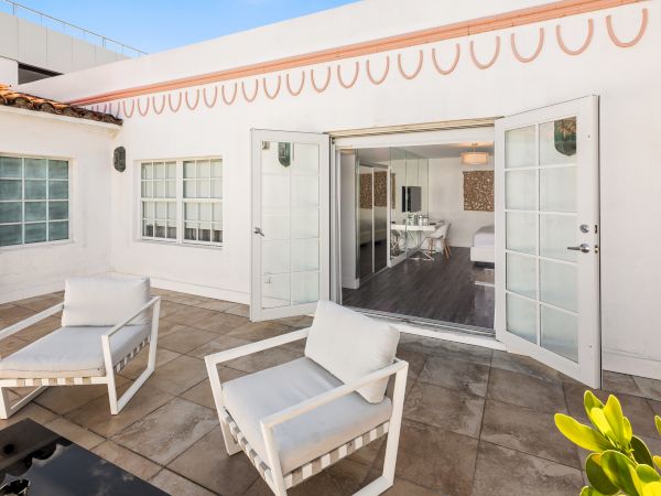 A sunlit patio features two white chairs, leading into a bright room through open double doors in a modern white building.