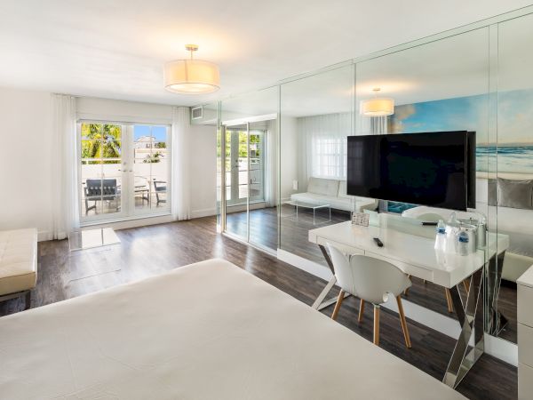 A modern hotel room with a bed, TV, desk, and mirrored wall, featuring a bright, airy design and an ocean-themed mural.