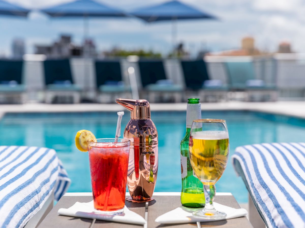 A poolside setup with two drinks, a cocktail shaker, and two striped towels. The background shows sun loungers and parasols by the pool.