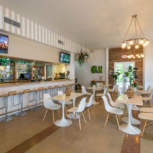 A modern and bright café/bar with white tables and chairs, a bar counter with stools, plants, and flat-screen TVs on the wall, ending the sentence.