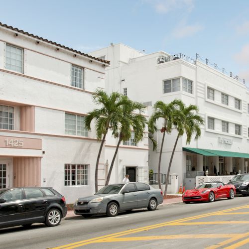 A street scene with white buildings, palm trees, and parked cars, including a red sports car, on a clear day with a few clouds in the sky.