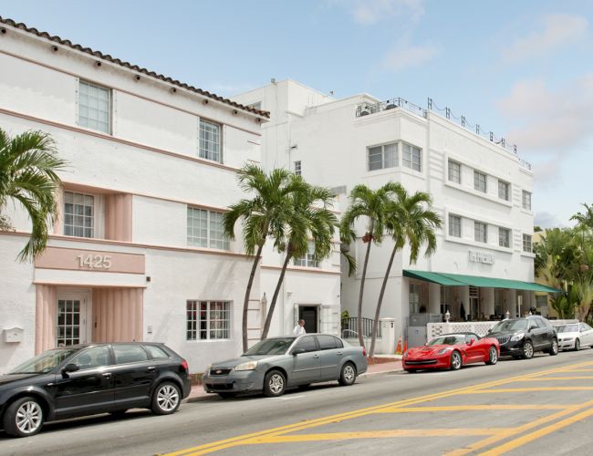 A street scene with white buildings, palm trees, and parked cars, including a red sports car, on a clear day with a few clouds in the sky.