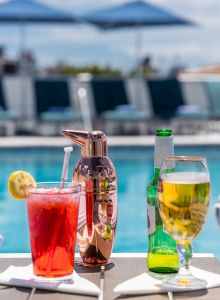 A poolside setting with striped towels, a red cocktail with lemon, a shaker, and a beer bottle with glass, with blue umbrellas and cityscape in the background.