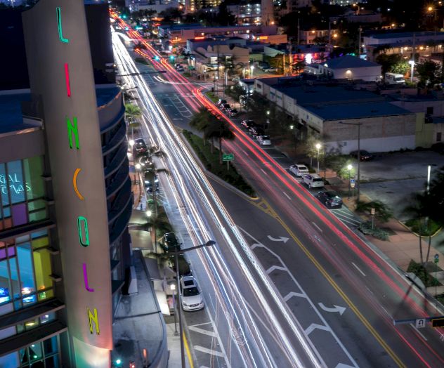 A cityscape at night featuring light trails from traffic on a busy street, tall buildings in the background, and a multi-colored sign on the left.