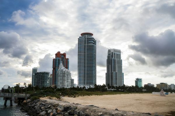 Tall buildings with a cloudy sky in the background, next to a rocky shoreline and sandy beach area.