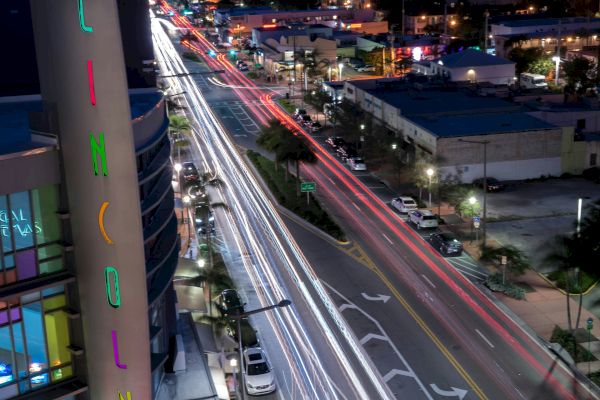 Cityscape at night featuring light trails from cars on a busy street, illuminated sign on a building, and high-rise buildings in the background.