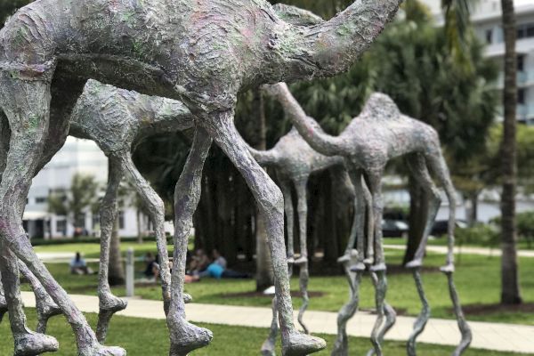 Sculptures of tall, abstract creatures with slender legs are displayed outdoors, set against a backdrop of palm trees and a grassy area.