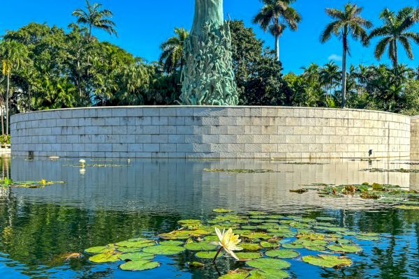 A sculpture rises from a circular stone platform surrounded by a water pond with lily pads, set against a backdrop of palm trees and clear blue sky.