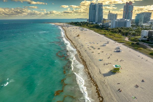 Aerial view of a beach with turquoise water, sandy shores, people, and tall buildings under a partly cloudy sky, possibly a coastal city.