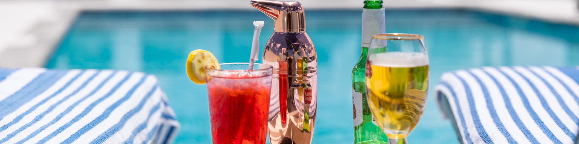 Two striped towels on lounge chairs by a pool, with a red drink, a beer, and a cocktail shaker on a small table, under blue umbrellas.